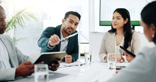 Ein Mann präsentiert während eines Meetings, während drei weitere Personen aufmerksam zuhören. Grafiken sind im Hintergrund sichtbar.