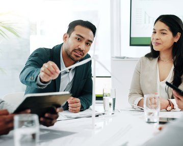 A man is presenting during a meeting while three other people listen attentively. Graphics are visible in the background.