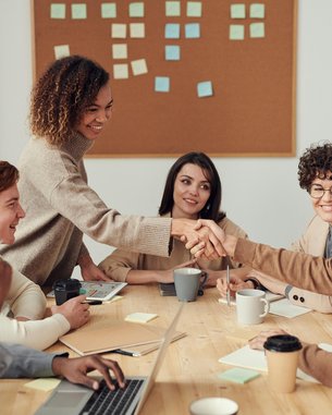 Sechs Personen in einem Besprechungsraum, die an einem Tisch sitzen. Zwei Personen schütteln sich die Hände, während andere zuschauen. Auf dem Tisch befinden sich Laptops, Tassen und Notizen. Ein Korkbrett im Hintergrund zeigt bunte Haftnotizen.