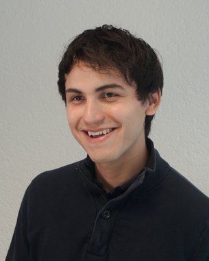 Ein junger Mann mit dunklen Haaren lächelt freundlich, trägt einen dunkelblauen Pullover und steht vor einer hellen Wand.