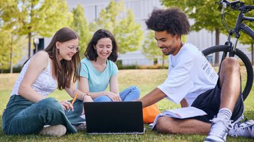 Three students are sitting on the grass, working together on a laptop. In the background, there is a bicycle.