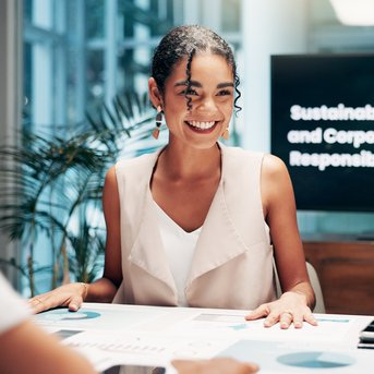 Two women are sitting at a table discussing. In the background, there is a screen displaying the text 'Sustainability and Corporate Responsibility.