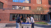Fünf Personen spielen Tischfußball im Freien, während zwei weitere Personen durch ein Fenster zuschauen.