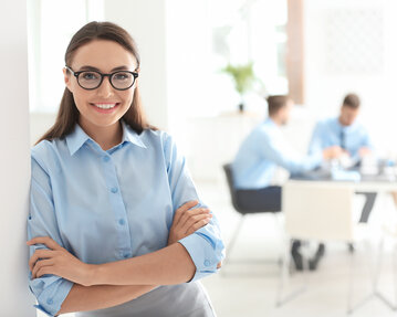 Eine Frau mit Brille und blauer Bluse steht lächelnd an einer Wand, während im Hintergrund eine Besprechung stattfindet.