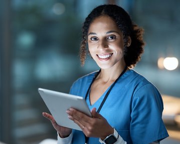 A nurse in a blue uniform is holding a tablet and smiling as she stands in a modern, well-lit room.