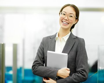 Eine lächelnde Frau in einem grauen Anzug hält ein Tablet in den Händen und steht in einem modernen Büro.