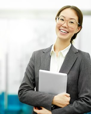 Eine lächelnde Frau in einem grauen Anzug hält ein Tablet in den Händen und steht in einem modernen Büro.