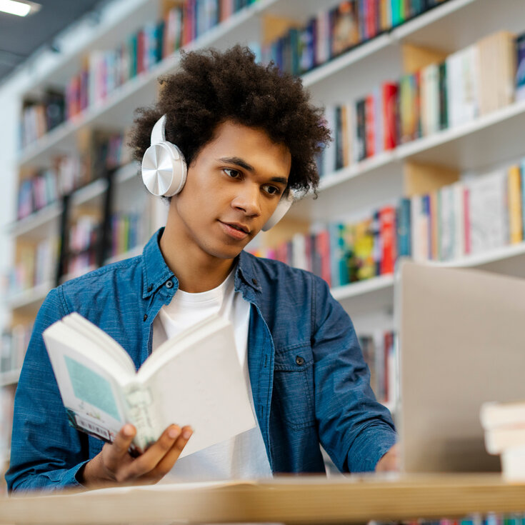 Ein junger Mann mit Kopfhörern liest ein Buch, während er an einem Laptop in einer Bibliothek sitzt, umgeben von Regalen voller Bücher.