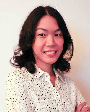 Eine Frau mit lockigem Haar trägt ein gepunktetes Hemd und steht mit verschränkten Armen vor einem neutralen Hintergrund.