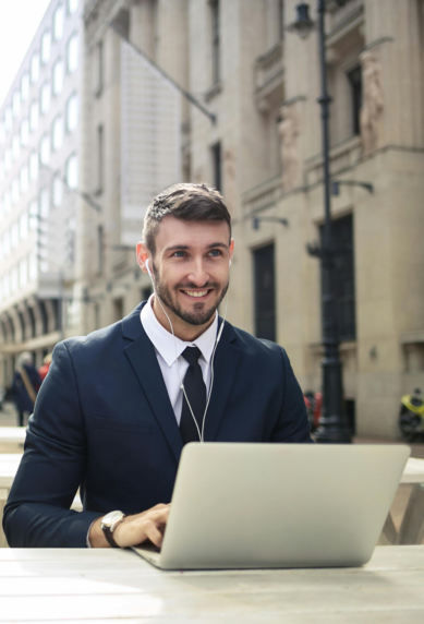 A man in a suit sits at an outdoor table, working on a laptop with a coffee beside him.