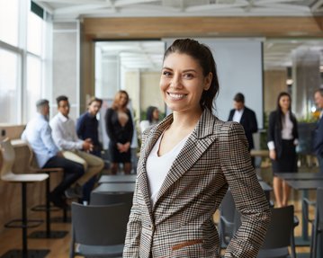 A smiling woman in a checked blazer stands in the foreground of a modern conference room, while several people are talking to each other in the background.