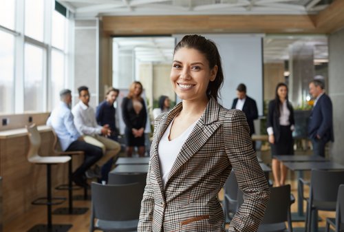 Eine lächelnde Frau in einem karierten Blazer steht im Vordergrund eines modernen Konferenzraums, während im Hintergrund mehrere Personen miteinander sprechen.