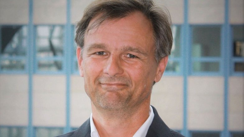 Ein Mann mit kurzen, grauen Haaren und einem leichten Lächeln trägt einen dunklen Anzug und steht vor einem modernen Hintergrund.