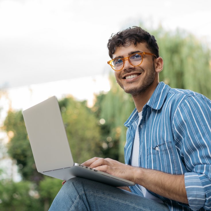 A young man with glasses is sitting outside, working on a laptop, smiling and relaxed.