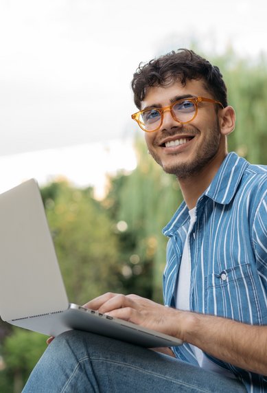 A young man with glasses is sitting outside, working on a laptop, smiling and relaxed.