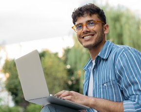 A young man with glasses is sitting outside, working on a laptop, smiling and relaxed.