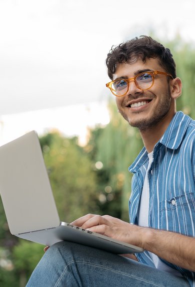 A young man with glasses is sitting outside, working on a laptop, smiling and relaxed.