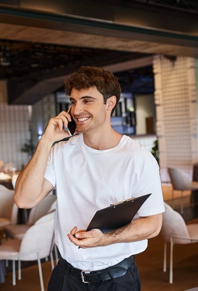 Ein junger Mann spricht am Telefon und hält einen Notizblock in einem elegant dekorierten Restaurant mit gedeckten Tischen.
