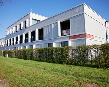 Moderne, mehrstöckige Gebäude mit weißen Wänden und großen Fenstern, umgeben von einer grünen Hecke. An der Fassade befindet sich ein Werbeschild der SRH.