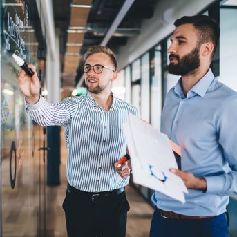 Two men in a modern office, one writing on a glass wall while the other holds notes and watches.