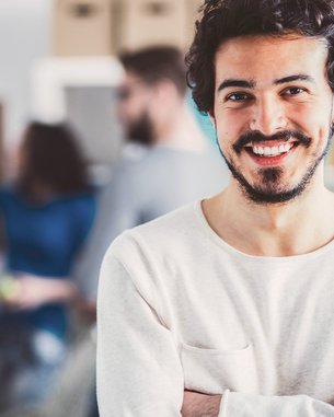 Ein junger Mann mit einem Bart lächelt und steht mit verschränkten Armen im Vordergrund. Im Hintergrund sind mehrere Personen zu sehen, die an einer kreativen Aktivität teilnehmen. Die Umgebung wirkt modern und einladend.