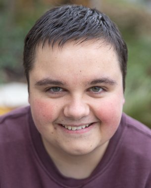 Ein junger Mann mit kurzen, dunklen Haaren und einem freundlichen Lächeln, trägt ein lila Sweatshirt und schaut direkt in die Kamera.