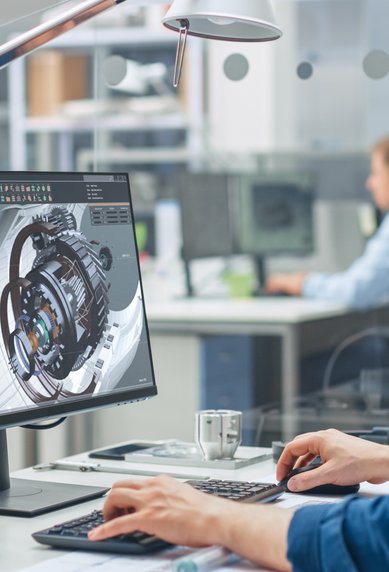A man is working at a computer that displays a technical design. In the background, other people can be seen in a modern office.