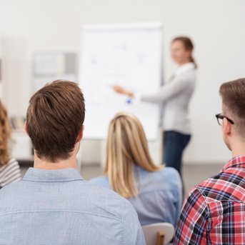 A group of people is sitting with their backs to the camera, listening to a presentation at a flip chart.