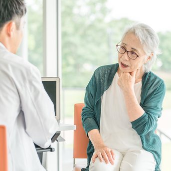 An older woman with glasses is sitting in a conversation with a doctor, appearing thoughtful. The doctor is seen from behind.