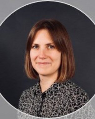 Eine Person mit schulterlangen, braunen Haaren und einem lächelnden Gesicht, trägt ein gemustertes Hemd und steht vor einem neutralen Hintergrund.