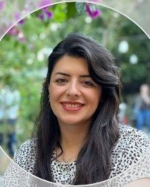 Eine Frau mit langen, dunklen Haaren und einem lächelnden Gesicht, gekleidet in einem weißen Oberteil mit schwarzem Muster, vor einer grünen, blühenden Kulisse.