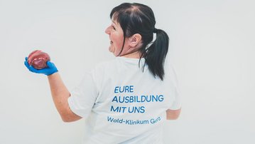 Eine Person mit einem weißen T-Shirt hält ein rotes Objekt in der Hand und zeigt auf den Rücken des Shirts mit dem Text „EURE AUSBILDUNG MIT UNS“.