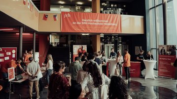 Menschen versammeln sich in der SRH Hochschule Heidelberg, um Informationen zu erhalten. An einem Empfangstisch stehen Mitarbeiter bereit, während ein Willkommensbanner im Hintergrund hängt. Die Atmosphäre ist geschäftig und einladend.