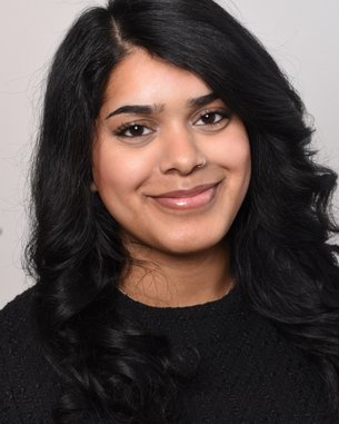 Porträt einer Frau mit langen, lockigen, schwarzen Haaren und einem freundlichen Lächeln, die einen schwarzen Pullover trägt.