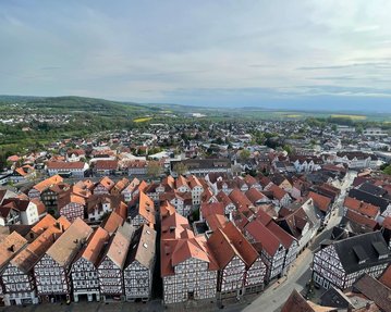 Blick auf eine Stadt mit Fachwerkhäusern und roten Dächern, umgeben von grünen Hügeln und Feldern im Hintergrund.