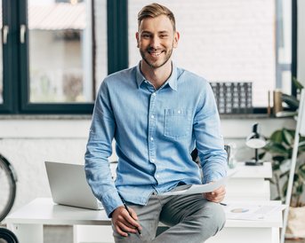 A man is sitting at a desk with a smile, holding a sheet of paper in one hand, with a laptop and a bicycle in the background.
