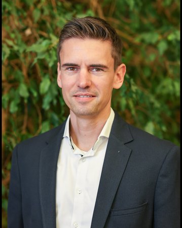 Ein Mann mit kurzen, braunen Haaren trägt einen dunklen Anzug und ein helles Hemd, vor einem Hintergrund aus grünen Pflanzen.