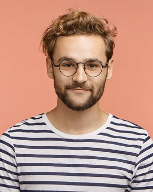 Junger Mann mit lockigem, blondem Haar und Brille, trägt ein gestreiftes T-Shirt vor einem einfarbigen, rosa Hintergrund.