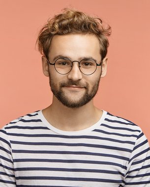 Junger Mann mit lockigem, blondem Haar und Brille, trägt ein gestreiftes T-Shirt vor einem einfarbigen, rosa Hintergrund.