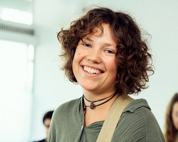 Lächelnde junge Frau mit lockigem Haar, die eine Gitarre hält. Im Hintergrund sind weitere Personen unscharf zu sehen.