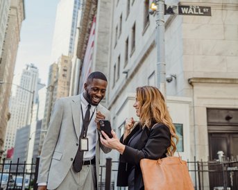 Zwei Personen stehen vor einem Gebäude an der Wall Street. Der Mann in einem Anzug lächelt, während die Frau mit einer Handtasche auf ihrem Smartphone schaut.