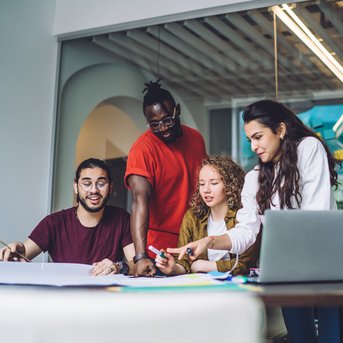 Eine Gruppe von vier Personen arbeitet gemeinsam an einem Tisch, umgeben von Notizen und einem Laptop. Sie diskutieren und zeichnen auf einem großen Blatt Papier.