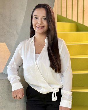A person with long brown hair is wearing a white blouse and black pants. They are standing by a wall and smiling. In the background, yellow steps are visible.