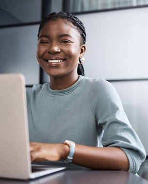 Eine lächelnde Frau mit Zopf sitzt an einem Tisch und arbeitet an einem Laptop in einem modernen Büro.