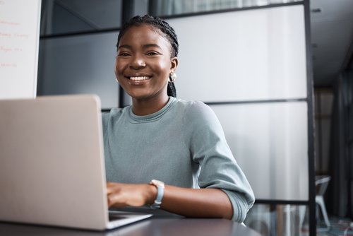 Eine lächelnde Frau mit Zopf sitzt an einem Tisch und arbeitet an einem Laptop in einem modernen Büro.
