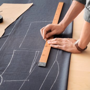 A hand is drawing with a ruler on black fabric to create a pattern. Next to the fabric are measuring tapes and scissors.