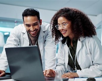 Zwei Wissenschaftler in Laborkitteln arbeiten gemeinsam an einem Laptop und schauen lächelnd auf den Bildschirm.