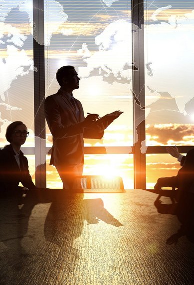 A business meeting with four people sitting at a table. One person is standing and presenting, while a world map and a sunset are visible in the background.
