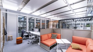 Moderne Büroumgebung mit orangefarbenen Sitzmöbeln, einem Tisch und Arbeitsplätzen. Große Fenster sorgen für viel Tageslicht. Der Raum ist funktional und einladend gestaltet.