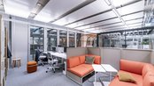 Moderne Büroumgebung mit orangefarbenen Sitzmöbeln, einem Tisch und Arbeitsplätzen. Große Fenster sorgen für viel Tageslicht. Der Raum ist funktional und einladend gestaltet.